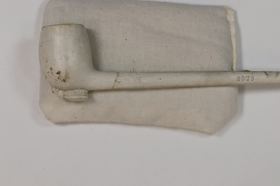 Hearst Museum object 2 of 6 titled Pipe w/ box, accession number 7-9664a-c, described as Pipe with box. (a) Pipe, white clay, long stem, foot with full relief of a bee, incised “Gambie a Paris m * M Depose 1079;” broken. Length 20 cm. Diameter of bowl 2.5 cm. (b), (c) Box, two pieces, wood, with brass catch, stained, broken. Length 22 cm. Diameter of bowl 3 cm.