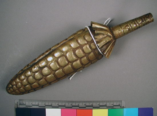 Hearst Museum object titled Corn figurine, accession number 16-20431, described as Chimu style corn. Repoussed, brass ear at corn, formed in 2 pieces then joined.  handle and end of shucks added.  small worm applied to one surface.  hollow.