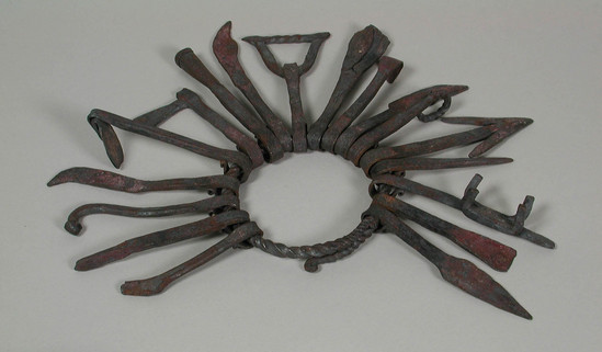 Hearst Museum object titled Bracelet, accession number 5-15671, described as Bracelet associated with the worship of Ogun (the god of blacksmiths). Wrought iron; twisted iron rod with overlapping opening; 18 miniature tools (hoe, axe, knife, etc) looped around ring; diameter ca. 7.5 cm; maximum length of tools 9.0 cm. Miniature tools include:  spear point(?), chisel(?), door lock, auger(?), arrowhead(?), ring, machete, calabash scraper, gong, stirrup or bow, knife(?), bell (aja), axe, hoe, knife, drumstick, half a tongs(?), hammer(?).