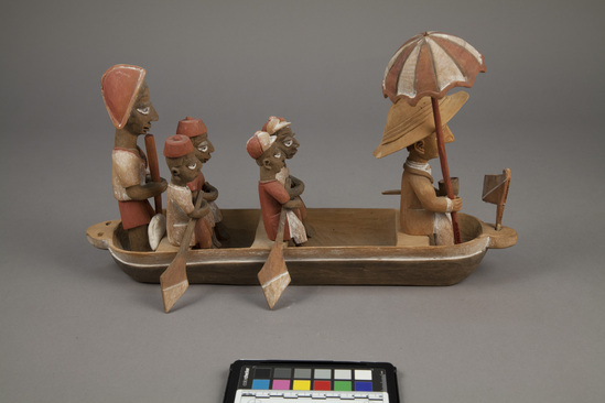 Hearst Museum object 14 of 15 titled Figures, accession number 5-16237a-q, described as caricature figure group; caricature of a District Officer on tour; carved & painted wood a - boat, has bag of frags b - Dist. officer (front) c - poler (rear) d - paddler (LF) e - paddler (LR) f - paddler (RF) g - paddler (RR) h - hat i - umbrella handle j - umbrella k - flag l - pipe m - pole n - paddle (RF) o - paddle (LF) p - paddle (LR) q - paddle (RR) Found two pegs in a bag with this object number.