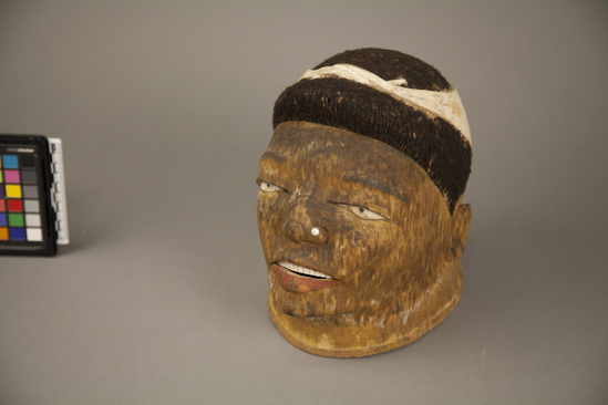 Hearst Museum object titled Mask, accession number 5-12996, described as helmet mask; anthropomorphic wood mask; open lips w/ red pigment on lower lip; metal stud through left nostril; whitened teeth and eyes; yellow ground; black human hair on head; head ornament of whitened carved wood w/ floral motif; split and repaired on lower right; h. 22.5 cm.