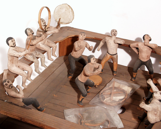 Hearst Museum object titled House model, accession number 2-4571, described as Model of interior of dance house. Square. 12 individually carved and painted men including 3 drummers seated on bench. All drums have gut heads and are broken. Made with wood and pigment.