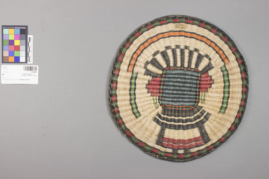 Hearst Museum object 2 of 3 titled Plaque, accession number 2-68500, described as Round, design showing kachina mask using black, red, green and orange on whitened, natural ground. Wicker weave, sumac warp, dyed rabbit brush weft, yucca for wrapped rim.