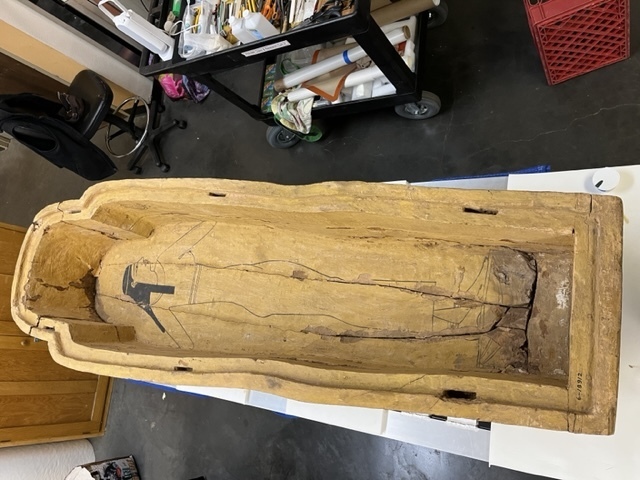 Hearst Museum object 37 of 167 titled Coffin, accession number 6-19912, described as Wooden anthropoid coffin. Inscription running down center of lid. Determined by Kea Johnston to probably be the outer coffin of Iwefaa (6-19928, 12-11039). According to Kea Johnston, "This coffin belonged to a man named Iwefaa, whose name is inserted in the middle of the spells written in the two columns on the front. This coffin is likely the outer coffin for PAHMA 6-19928 based on the similarity of the owner's name, unusual costume, and the paleography of the inscription. Both coffins were purchased on the antiquities market by George Reisner for Phoebe A Hearst. However, stylistically, they are very similar to a set of coffins known to have come from the site of Akhmim, which was excavated in the 1880s by Gaston Maspero, and which has heavily looted for the antiquities market during this decade." "The pectoral vignette between the text and the collar shows the deceased standing before squatting baboon and is the source of our logo. The hieroglyph for 'sky' is above their heads, and the side borders are formed by a pyramid and the symbol for the West.