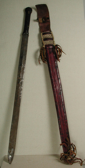 Hearst Museum object titled Sword and sheath, accession number 5-11719a,b, described as sword and sheath;  metal blade with dark brown leather covered handle, red leather sheath with faint black design, red leather fringe and black and white plaited leather strips decorate sheath;  71 cm long.