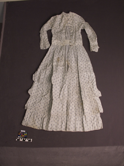 Hearst Museum object titled Dress, accession number 2-66958, described as Lightweight, white cotton dress with all-over small floral print in blue and black, long sleeves, tired ruffled skirt and an opening in the back with mother-of-pearl buttons.  Garment is age-spotted.