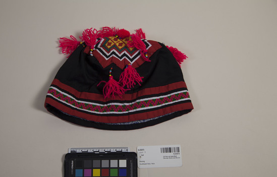 Hearst Museum object titled Hat, accession number 9-23373, described as Hmong baby hat Black hat with bottom band of horizontal stripes in appliqué and cross stitch. Top appliqué, yarn pompom, yarn tassels hanging from strings of beads.