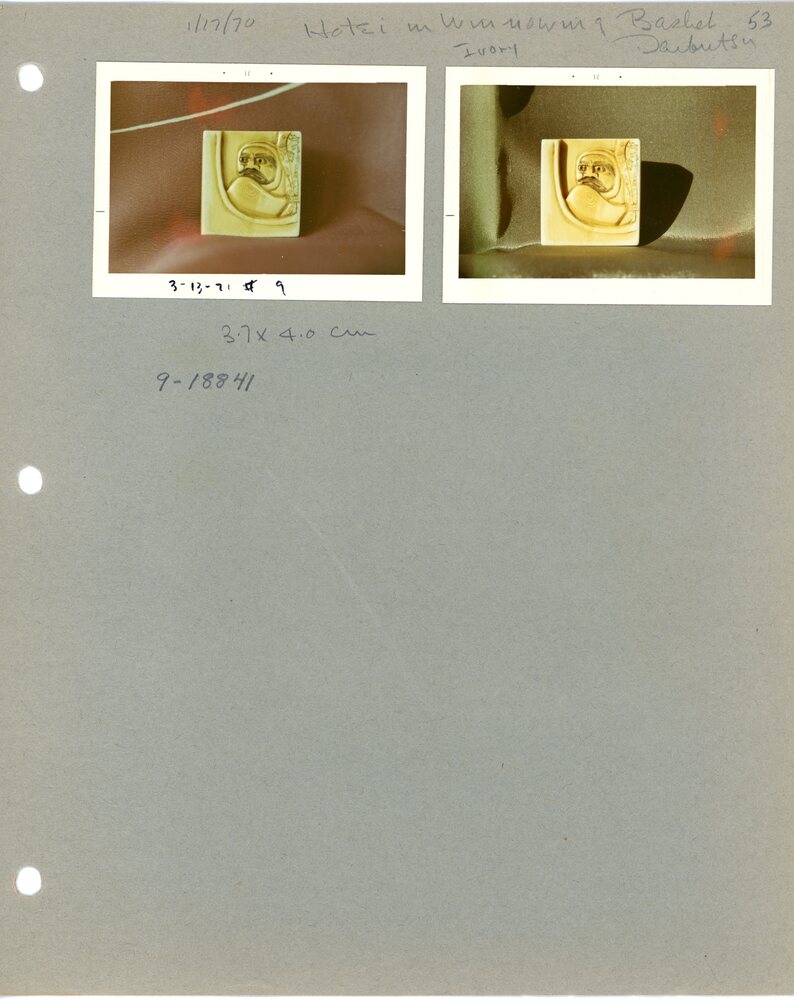 Hearst Museum object 5 of 7 titled Netsuke, accession number 9-18841, described as Netsuke; "Daruma and treasures in winnowing basket;" rectangular, with four ridge lines as on a roof on one side; head of a man in low relief, cloak held up to chin; eyes and mouth painted; "curtain" with incised objects to his left; L 4.0 cm X W 3.6 cm