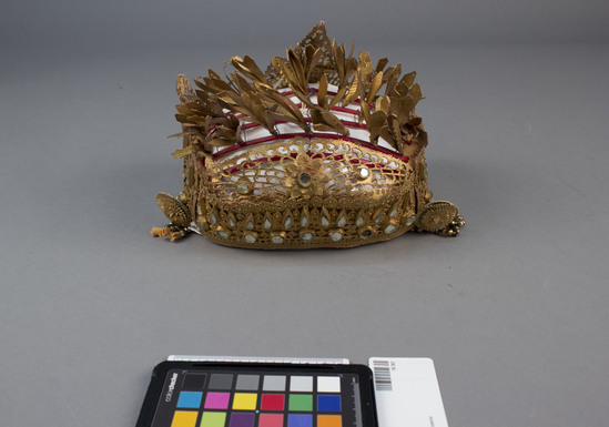 Hearst Museum object titled Hat, accession number 18-367, described as Gold-leafed leather headdress; glass ornaments. Headdress worn filled with flowers from “Cambodia” tree. Width 8 inches. 18- 367 - 379 comprise young girl’s festival and “Legong” dance costume; see photocopy of page from Covarrubias, “Island of Bali.”