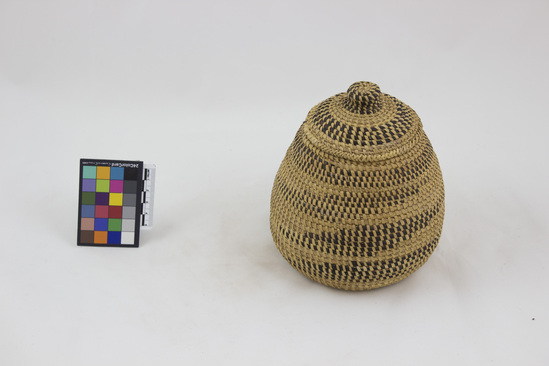 Hearst Museum object titled Basket, accession number 5-6722a,b, described as basket with lid; grass in coiling technique on bundle core; wide stitching, both inter & interlocking type; natural color with black designs; herring bone over-stitch rims;  a) basket; sub-globular shape with bulging base; b) lid; flattened beehive shape with knob;  overall height 17.5 cm; diameter base 13 cm. Native name: Ekigega. Used by men and women for storage. Collected July 1967.