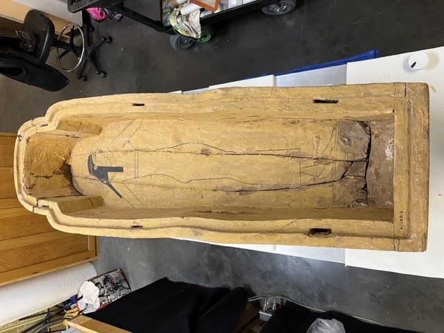 Hearst Museum object 69 of 167 titled Coffin, accession number 6-19912, described as Wooden anthropoid coffin. Inscription running down center of lid. Determined by Kea Johnston to probably be the outer coffin of Iwefaa (6-19928, 12-11039). According to Kea Johnston, "This coffin belonged to a man named Iwefaa, whose name is inserted in the middle of the spells written in the two columns on the front. This coffin is likely the outer coffin for PAHMA 6-19928 based on the similarity of the owner's name, unusual costume, and the paleography of the inscription. Both coffins were purchased on the antiquities market by George Reisner for Phoebe A Hearst. However, stylistically, they are very similar to a set of coffins known to have come from the site of Akhmim, which was excavated in the 1880s by Gaston Maspero, and which has heavily looted for the antiquities market during this decade." "The pectoral vignette between the text and the collar shows the deceased standing before squatting baboon and is the source of our logo. The hieroglyph for 'sky' is above their heads, and the side borders are formed by a pyramid and the symbol for the West.