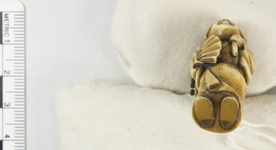 Hearst Museum object titled Netsuke, accession number 9-18774, described as Netsuke; figure of standing woman carrying open fan in right hand; carrying rectangular box on back, supported by cord passing through ring on top of box, and held in her left hand; four drawers opening on side of box; leaf incised on back of box; H 4.2 cm X W 1.3 cm