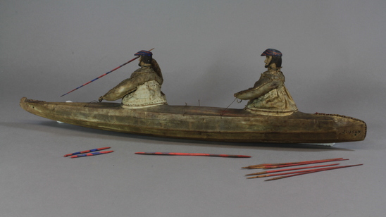 Hearst Museum object 3 of 8 titled Kayak model, accession number 2-4569, described as 2-hatch. Wood frame, rawhide cover. 2 human figures wearing hats painted bright blue and red and wearing sealskin parkas. 15 fixtures including weapons and paddles painted bright red and blue.