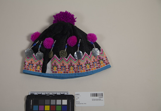 Hearst Museum object titled Hat, accession number 9-23372, described as Hmong baby hat, black velvet pompoms, embroidery, metal decorations, beads. Lined. Body of had made with factory patterned textile. Band of appliqué with cross stitching over, yarn pompoms, strings of plastic and metal beads ending in metal tabs.