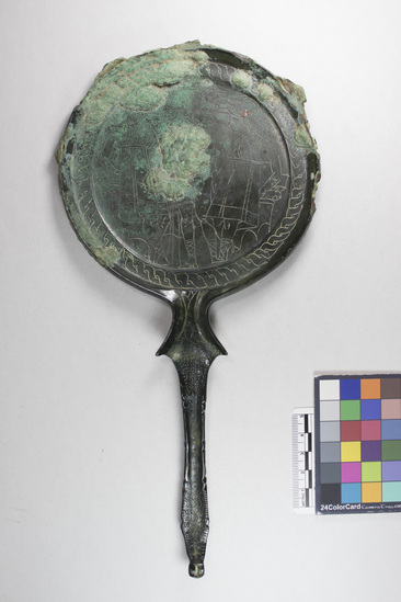 Hearst Museum object 6 of 13 titled Stemmed mirror, accession number 8-5859, described as Etruscan bronze mirror; cast-handle type; encrustation on both surfaces of disk and blue discoloration on obverse due to bronze disease; dark green patina; handle ends in stylized ram’s head; rim of obverse beaded; engraved scene of four figures, two women flanking Minerva (Greek=Athena) and nude man, standing against architecture; guilloche border; mirror appears to have been conserved with a translucent glaze.