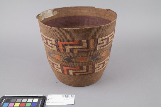 Hearst Museum object 2 of 3 titled Basket, accession number 2-30995, described as Cylindrical, twined spruce root, exterior false embroidery of grass stems dyed yellow, orange, black and blue; central chevron-like designs, flanked top and bottom by designs on maroon dyed spruce root bands. Rim slightly damaged; cardboard sewn to base. Broad maroon and narrow green and black dyed spruce root bands in interior.