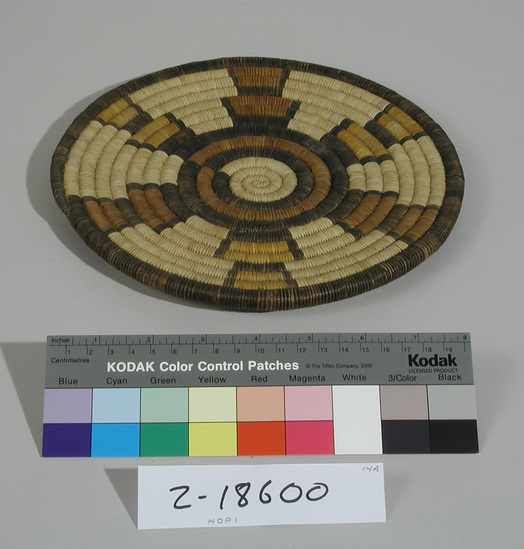 Hearst Museum object titled Plaque, accession number 2-18600, described as Basketry, coiled.  Shades of brown and yellow.  Star design:  6 arms radiate out to rim coil.  Around rim 2 stacked trapezoids with alternating light brown - yellow - light brown.  Rim coil is brown, alternating light/dark dyes.  Coiled loop handle.