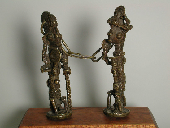 Hearst Museum object 2 of 10 titled Figures, accession number 5-16317, described as Ogboni society figures; pair of Ogboni society figures connected by a chain and mounted on a wooden block, cast brass figures, (a) 1 male, (b) 1 female, each holding a staff and rattle. both have elaborate hair treatment and head pieces