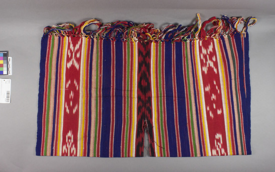 Hearst Museum object titled Shawl, accession number 16-20213, described as Child’s poncho; 66.5 centimeter w, 37 centimeter. l ,excluding fringe. 13 centimeter fringe. Multi colored small bands with three large bands of ikat design;11 centimeter vertical head opening, machine stitched. fringe on one length vertical warp tied fringe; fringe on other length separate  has separate bundles tied/looped onto cut warp ends of fabric