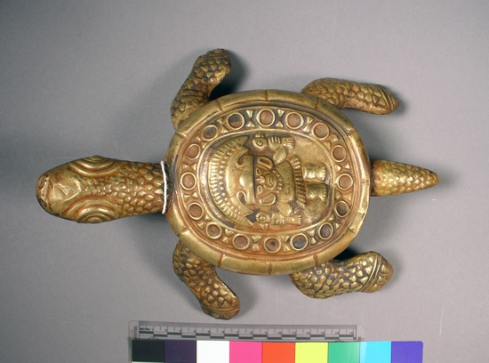 Hearst Museum object titled Turtle figurine, accession number 16-20402, described as Chimu style turtle of gold-colored metal. Male figure on back holding birds in each hand. Pattern of circles and diamonds in circular arrangement surrounding figure.