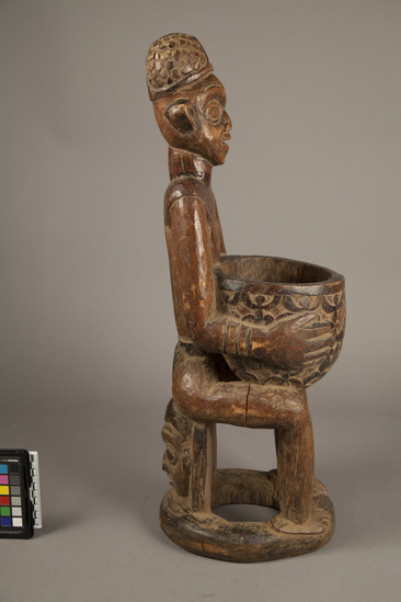 Hearst Museum object titled Carved wood figure, accession number 5-6001, described as Wood male figure seated on stool; holding bowl with human-like figures in low relief; stool is carved human head; h. 63.5 cm. Fine brown patina; some termite damage on base.