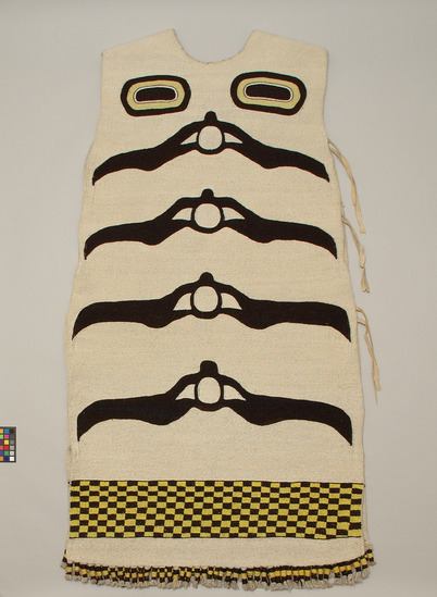 Hearst Museum object 2 of 3 titled Tunic, accession number 2-59550, described as Abstracted bear designs; black, yellow and blue on white; dyed mountain goat wool twined over wool-covered cedar bark warp; front design is standing bear with large face; smaller faces within ears and at joints; on back are (top to bottom) a pair of eyes, 4 spinal vertebrae?, and black/yellow checkerboard panel; tassels at base of garment; one side of tunic is sewn almost closed, other side has 3 pair buckskin ties; drawstring at neck. Designs said to be grizzly bear and 2 cubs.