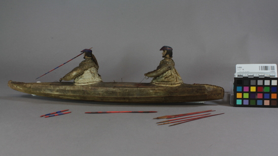 Hearst Museum object 7 of 8 titled Kayak model, accession number 2-4569, described as 2-hatch. Wood frame, rawhide cover. 2 human figures wearing hats painted bright blue and red and wearing sealskin parkas. 15 fixtures including weapons and paddles painted bright red and blue.