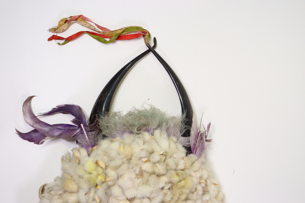 Hearst Museum object 15 of 15 titled Headdress, accession number 2-45410, described as Felt cap covered with strips of rabbit fur, patches stained yellow, traces of green fluff at front. Crown with 2 split polished horns attached to interior hide strip. Purple feathers and green-gray fluff in between and at base of horns. Multicolored silk grosgrain (?) ribbons bound to left horn tip. Ten "ermine tails" of rabbit fur sewn about a cord, red felt seized, with black hair tips; fringes at sides.