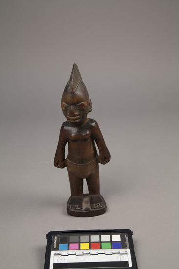 Hearst Museum object 5 of 6 titled Ere ibeji figure, accession number 5-15807, described as Figure, twin (Ibeji), wood, handcarved; female, with elongated headdress, wearing grass skirt; hands on hips; straight legs, with feet mounted on keystone-shaped base; dark brown, polished finish. Bottom stamped "Product of the Gold Coast" by William Bascom in compliance with customs regulation, per collector's notes.