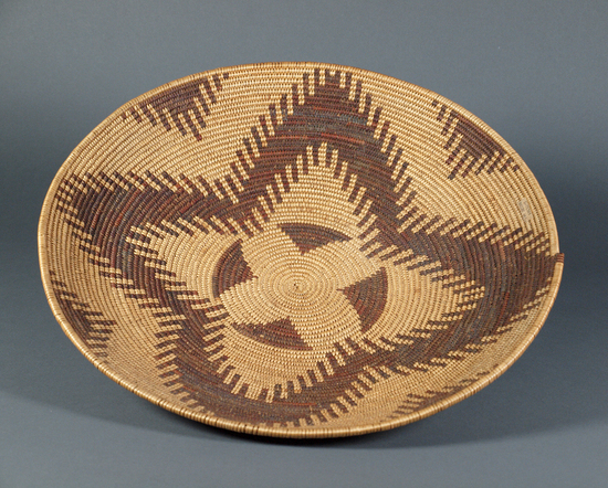 Hearst Museum object 3 of 4 titled Winnowing tray, accession number 1-71369, described as Coiled basket, deep tray.  Tag "Maidu att.".  Tag on baskets "D-62". Per Ralph Shanks:  Coiled basket winnowing tray.  Tight spiral, pinhole start.  The basket has a three rod foundation, probably willow.  The weft materials are peeled and unpeeled redbud.  The design at the base is a four sided star/flower.  On the sides is one horizontal band of zigzags, forming a four pointed star/flower.   The workface is on the inside, with many split stitches on the backface.  The weft fag ends are concealed and the work direction is to the left.  The basket has a plain wrapped rim with a blunt coil ending.  This is basket is attributed to the Patwin due to the shape and depth of the basket and the peeled redbud background.