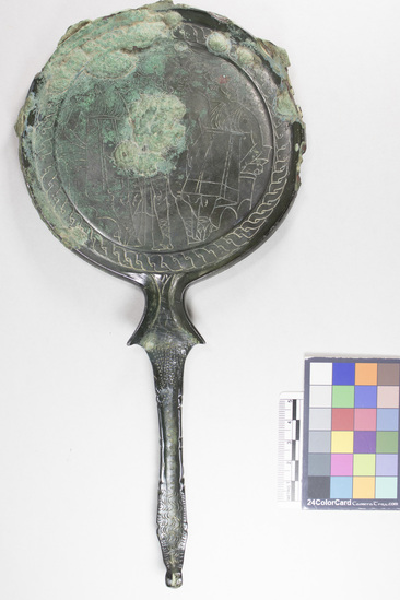 Hearst Museum object 12 of 13 titled Stemmed mirror, accession number 8-5859, described as Etruscan bronze mirror; cast-handle type; encrustation on both surfaces of disk and blue discoloration on obverse due to bronze disease; dark green patina; handle ends in stylized ram’s head; rim of obverse beaded; engraved scene of four figures, two women flanking Minerva (Greek=Athena) and nude man, standing against architecture; guilloche border; mirror appears to have been conserved with a translucent glaze.