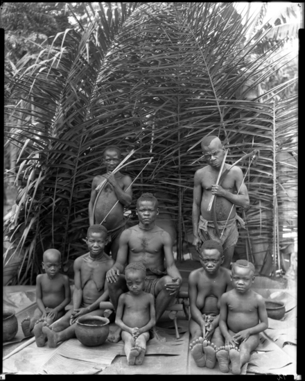 Hearst Museum object titled Black and white negative, accession number 15-32567, described as 8 x 10 " Negative. Image depicting a head chief, their family, and ministers, all posing for the camera. Hoefler's original description: "Belgian Congo. A royal court in pygmy-land. Beneath a canopy of palm and banana leaves the head-chief, or king, held court. His family and ministers attended upon him. In front of this throne in the clearing the drums beat and his subjects dances. The rhythm soon overpowered the royal dignity and it was not long until the whole court joined the dance.