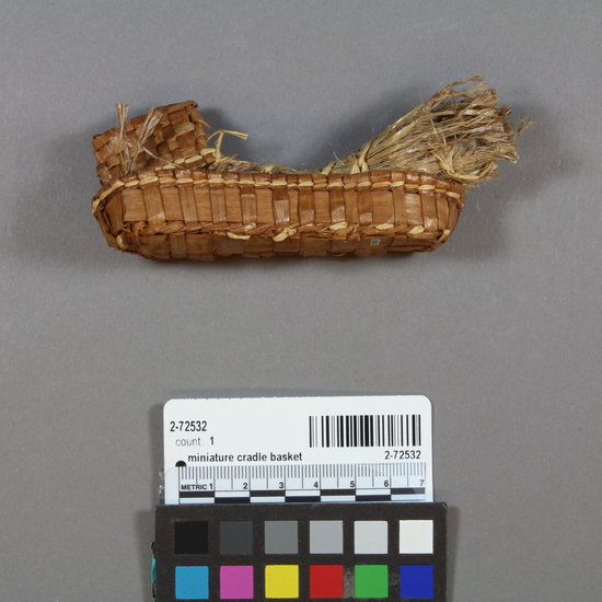 Hearst Museum object titled Miniature cradle basket, accession number 2-72532, described as Woven miniature cradle basket, made of cedar bark and shredded cedar bark, sewn and laced with raffia.  Baby "body" is still in place but head is missing. 1942.