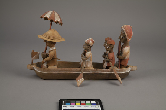 Hearst Museum object 10 of 15 titled Figures, accession number 5-16237a-q, described as caricature figure group; caricature of a District Officer on tour; carved & painted wood a - boat, has bag of frags b - Dist. officer (front) c - poler (rear) d - paddler (LF) e - paddler (LR) f - paddler (RF) g - paddler (RR) h - hat i - umbrella handle j - umbrella k - flag l - pipe m - pole n - paddle (RF) o - paddle (LF) p - paddle (LR) q - paddle (RR) Found two pegs in a bag with this object number.