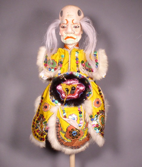 Hearst Museum object 5 of 6 titled Hand puppet, accession number 9-22437, described as Puppet, hand.  A.  Head:  carved wood, off-white, red mouth, pink cheeks, green facial hair, brown and black eyes,  2 tufts of long, white hair.  B.  Body:  cloth, white.  Legs:  cloth, tan.  Hands:  wood, white, orange nails.  Boots:  wood, black and white.  C.  Robe:  cloth, yellow with green, blue, pink and silver sequins, metallic pink animals face on front, black and white rabbit fur trim.  47 x 26 cm (18 1/2 x 10 1/4 in)