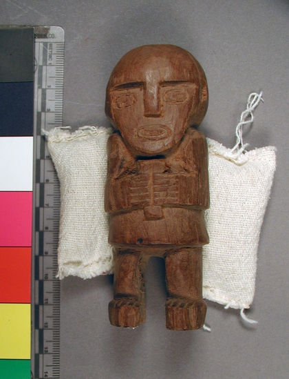 Hearst Museum object titled Figure, accession number 16-20437, described as carved wooden Chimu style figure. Static with large head, high shoulders, short kilt and stumpy legs over blocky feet. hands meet at center chest and are holding a small, unidentified object. carved features.