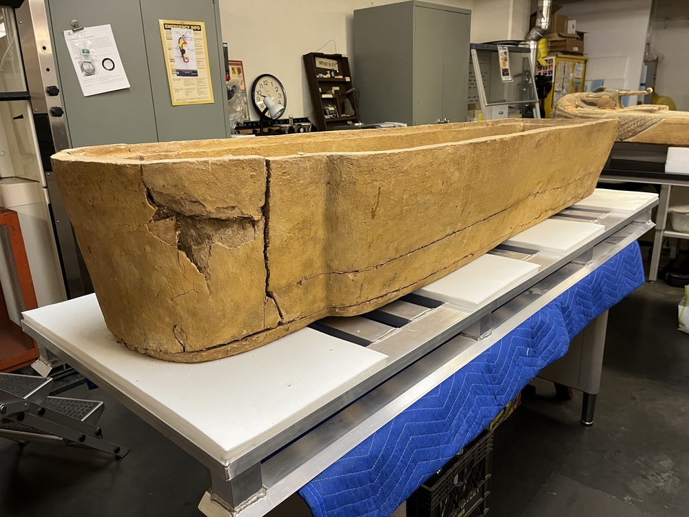 Hearst Museum object 166 of 167 titled Coffin, accession number 6-19912, described as Wooden anthropoid coffin. Inscription running down center of lid. Determined by Kea Johnston to probably be the outer coffin of Iwefaa (6-19928, 12-11039). According to Kea Johnston, "This coffin belonged to a man named Iwefaa, whose name is inserted in the middle of the spells written in the two columns on the front. This coffin is likely the outer coffin for PAHMA 6-19928 based on the similarity of the owner's name, unusual costume, and the paleography of the inscription. Both coffins were purchased on the antiquities market by George Reisner for Phoebe A Hearst. However, stylistically, they are very similar to a set of coffins known to have come from the site of Akhmim, which was excavated in the 1880s by Gaston Maspero, and which has heavily looted for the antiquities market during this decade." "The pectoral vignette between the text and the collar shows the deceased standing before squatting baboon and is the source of our logo. The hieroglyph for 'sky' is above their heads, and the side borders are formed by a pyramid and the symbol for the West.