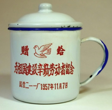 Hearst Museum object titled Mug with lid, accession number 9-23906a,b, described as Enamel mug with lid. The white ground is stenciled with a red inscription arranged in three lines. The top line, consisting of two characters “gifting to” (贈给zenggei), is inserted with a dove in the middle. The rest of the two lines states that it was specially made in commemoration of “the hardworking labors for the cause of constructing motherland” by State-operated 211 Factory in 7th November, 1957. Cobalt blue bowstring decorates the rim. The handle is partially decorated in the same color. RMB 100