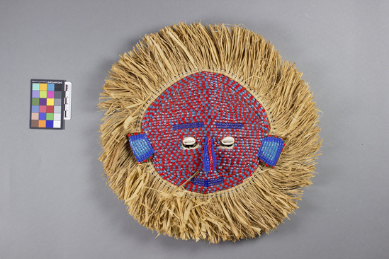 Hearst Museum object titled Mask, accession number 5-13760, described as Mask; face of short tubular beads over reed and cloth framework; beads alternating red and light blue, eyebrows, nose and mouth dark blue; cowrie shell eyes above eye slits; ears two shades of blue with red/green border; raffia edging about whole mask; ca. 35 cm