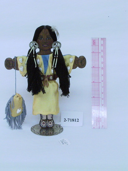 Hearst Museum object titled Doll, accession number 2-71812, described as Female, Osage doll.  Flat wooden doll with painted features and moccasins.  Dressed in golden silk dress decorated with cowrie shells and blue painted neck designs [front and back].  Thread hair with silver “conchos” and danglers at each side of face.  Feather in hair.  Leather belt with silver balls.  Matching silk bag holding sage from South Dakota.  Standing on a flat silver base.  H = 6.5”.