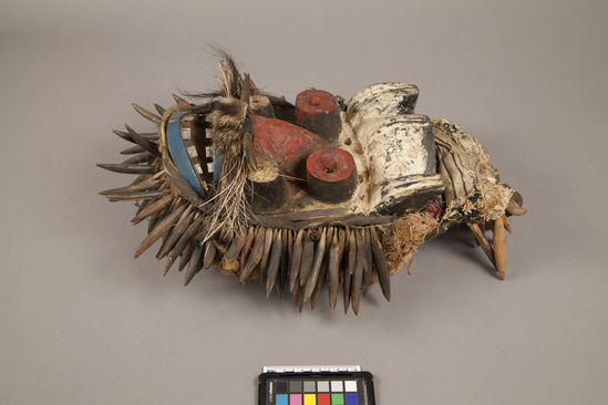 Hearst Museum object titled Mask, accession number 5-16217, described as carved and painted wood--red, white, blue, black, with wooden spike and vegetal material ruff. movable headpiece with wood and horn spikes and bells attached. heavy blue/white fabric. animal whisker mustache, metal teeth. small nails protrude randomly around raised, tubular eyes. back pierce for fabric hood to be attached. Purchased from Ron Atwood
