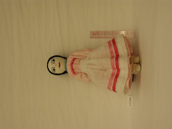 Hearst Museum object titled Female doll, accession number 2-71760, described as Southern Plains girl doll.   Cotton stuffed doll with embroidered features & black yarn hair.  Dressed in white suede mocassins, white cotton underpants and slip and apron & and a pink dress with pink and rose ribbon trim.  Apron trimmed with same ribbon as dress. Blue bead 6-strand necklace. Hair tied back into stiff, hourglass shaped bun near the top back of the head; decorated w/ long dangles of rose, green and pink ribbon with gold beads [a modern adaptation of typical young girls headdress for girls 8-10 years old].  H = 12”