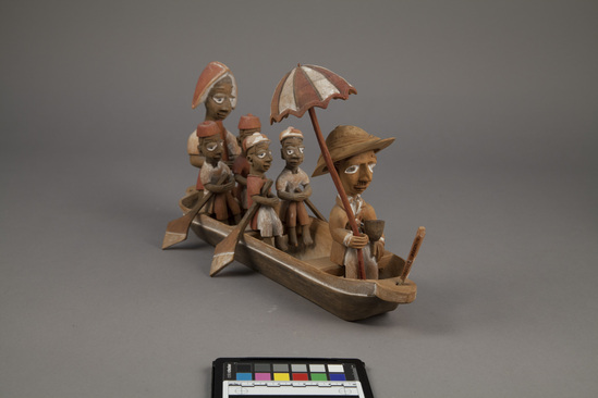 Hearst Museum object 13 of 15 titled Figures, accession number 5-16237a-q, described as caricature figure group; caricature of a District Officer on tour; carved & painted wood a - boat, has bag of frags b - Dist. officer (front) c - poler (rear) d - paddler (LF) e - paddler (LR) f - paddler (RF) g - paddler (RR) h - hat i - umbrella handle j - umbrella k - flag l - pipe m - pole n - paddle (RF) o - paddle (LF) p - paddle (LR) q - paddle (RR) Found two pegs in a bag with this object number.