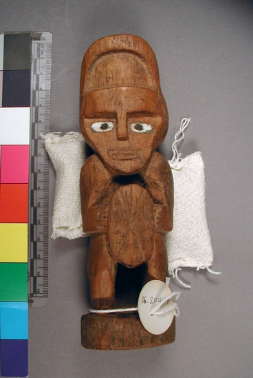 Hearst Museum object titled Figure, accession number 16-20444, described as Chimu style figure. Carved wooden figure with inlaid eyes (bone?). Figure wears a frontlet which stands 2.5cm above his brow, and holds an oval device (possibly a shield) in front of his body. figure is sitting on a very low stoop and holds the shield with both hands.