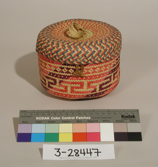 Hearst Museum object titled Basket and lid, accession number 3-28447a,b, described as Basket with lid; round, twill plaited palm leaf strips dyed purple, red and green, some left natural; lid is sewn to basket;  (a) basket: 2 rings sewn to base; diam. underside 16 cm; lid has loop at top to pull off; lid has leather closure which attaches to wood toggle on basket;  diam. lid ca. 16.7 cm