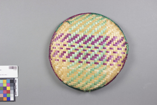 Hearst Museum object 1 of 2 titled Woven plate, accession number 5-11475, described as Woven plate, Nigeria, northern provinces, Hausa att. July 27, 1972. 5-11475, acc no. 2879, file code 1.5. Collector: T.D. Lanagan. Donor: W.H. Lawson. Materials, techniques: reeds split, some dyed; plaited into basket tray; rim added, consists of reed bundle wound about with reed strips Context of use: as container for household; Description: Basket tray, plaited; split reeds; white, purple, green; design of cross hatching in center; rim is alternating purple and green sections, raised; 25.5 cm. diameter; rim is 3 cm. h.;