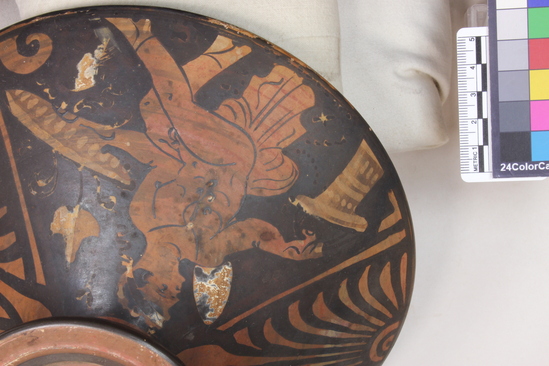 Hearst Museum object titled Lekanis lid, accession number 8-454, described as Apulian Red-figure lekanis lid; chips in rim; buff clay, reddened; knob in 2 degrees with central depression, decorated with rosette; A, male figure (fillet, petasos, sandals) seated on mantle, holding basket in r. hand, phiale in l. hand; B, woman seated on stool, holding casket in r. hand; between figures, palmettes; on rim, wave pattern.