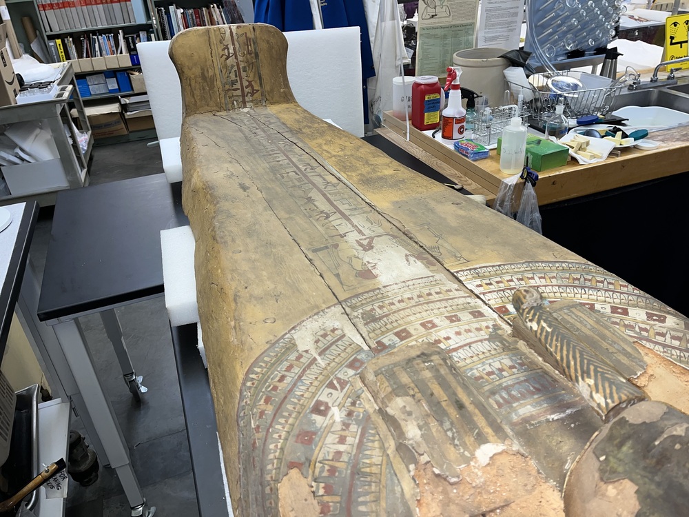 Hearst Museum object 66 of 167 titled Coffin, accession number 6-19912, described as Wooden anthropoid coffin. Inscription running down center of lid. Determined by Kea Johnston to probably be the outer coffin of Iwefaa (6-19928, 12-11039). According to Kea Johnston, "This coffin belonged to a man named Iwefaa, whose name is inserted in the middle of the spells written in the two columns on the front. This coffin is likely the outer coffin for PAHMA 6-19928 based on the similarity of the owner's name, unusual costume, and the paleography of the inscription. Both coffins were purchased on the antiquities market by George Reisner for Phoebe A Hearst. However, stylistically, they are very similar to a set of coffins known to have come from the site of Akhmim, which was excavated in the 1880s by Gaston Maspero, and which has heavily looted for the antiquities market during this decade." "The pectoral vignette between the text and the collar shows the deceased standing before squatting baboon and is the source of our logo. The hieroglyph for 'sky' is above their heads, and the side borders are formed by a pyramid and the symbol for the West.