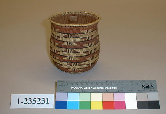 Hearst Museum object titled Basket, accession number 1-235231, described as Tall, narrow, flared fancy basket. 4 encircling geometric design bands in red with dark brown outlines.  There is a swastika in ink on the interior of the basket.  Tag "Kl. R. Tr. Att.". Per Ralph Shanks:  Twined globular basket vase, probably made for sale. Crossed warp starting knot.  The warp material is probably hazel. The weft material is conifer root.  The weft overlay design is beargrass, red dyed woodwardia, and maidenhair fern.  Starting at the starting knot, there is 1/2 inch of three strand twining, followed by 1 1/2 inches plain twining, followed by 2 weft rows of three strand twining.  Plain twining continues to the rim where there is one weft row of three strand twining followed by 1/4 inch of crossed warp open work.  The rim appears to be comprised of 1 weft row of twining with reinforcing rods wrapped with beargrass.  The main design is three horizontal rows of stepped parallelograms and triangles.  The basket has a rightward work direction, with up to the right slant of weft twist.  The workface is on the exterior.  The overlay is single-sided, on the exterior.  The basket is from Northwest California.  There is a possible human hair woven into the basket, near the center exterior.