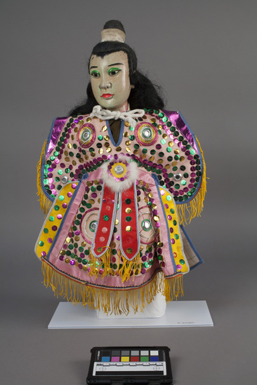 Hearst Museum object 8 of 10 titled Hand puppet, accession number 9-22260, described as Puppet, hand.  Young Man.  A.  Head:  wood, carved, white and pink with green, pink and black features, black hair in bun.  B.  Body:  cloth, brown.  Legs:  cloth, green and pink.  Hands:  wood, white.  Boots:  wood, black and white.  C. Robe:  cloth, pink with green, pink and gold sequins.  yellow fringe.  50 x 36 cm (19 11/16 x 14 3/16 in).